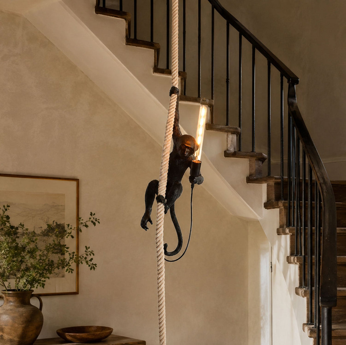 A black monkey pendant from Clérachic glows with a long filament bulb, hanging on its rope beside a staircase with a dark iron balustrade.