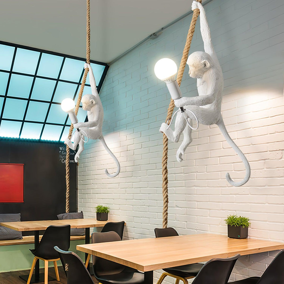 Two white monkey pendants on ropes with round bulbs, hung in a dining space with a white brick wall and pale wood tables, from Clérachic.