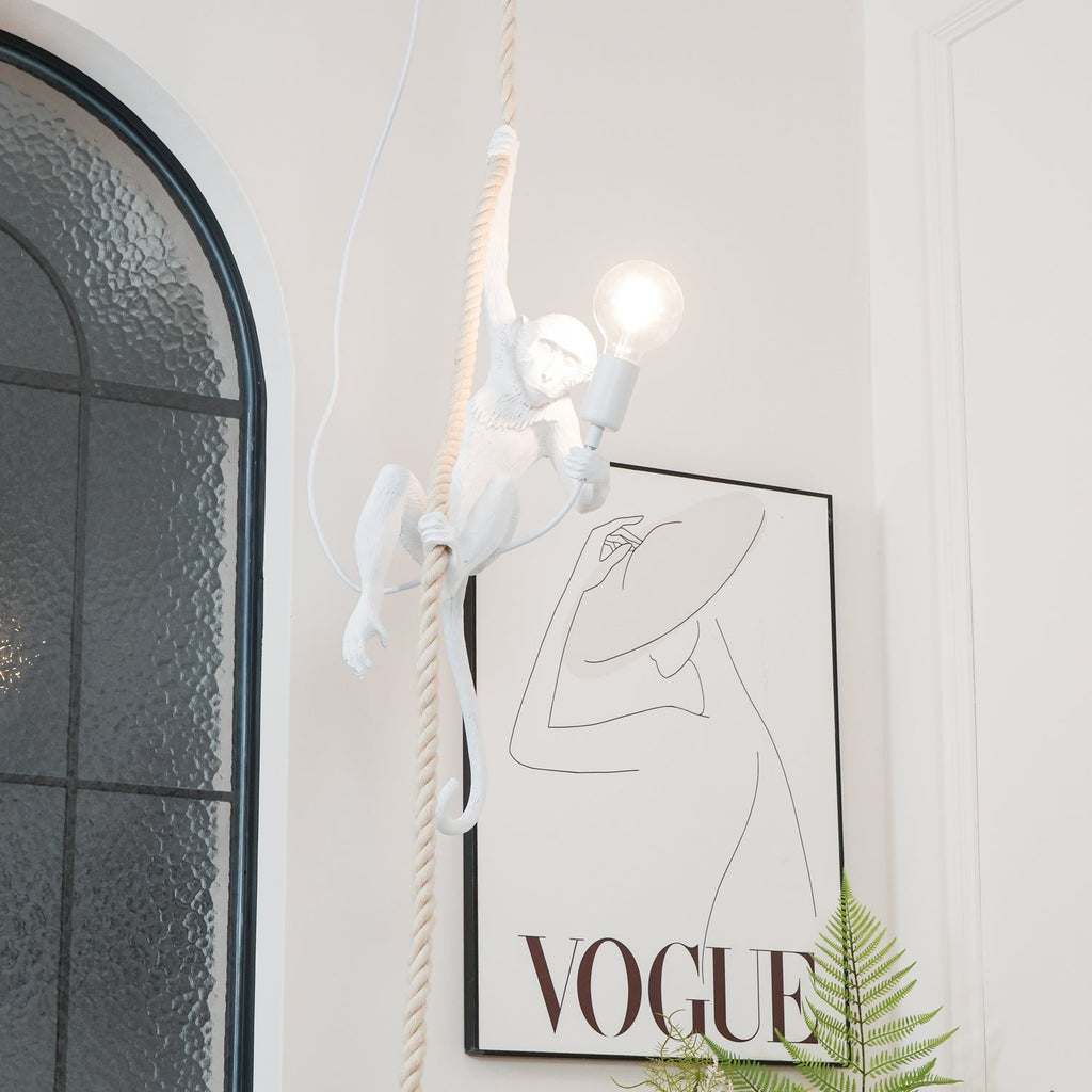 White resin monkey from Clérachic reaching along its rope with a glowing bulb, beside an arched glass window and a line-drawing print.