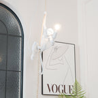White resin monkey from Clérachic reaching along its rope with a glowing bulb, beside an arched glass window and a line-drawing print.