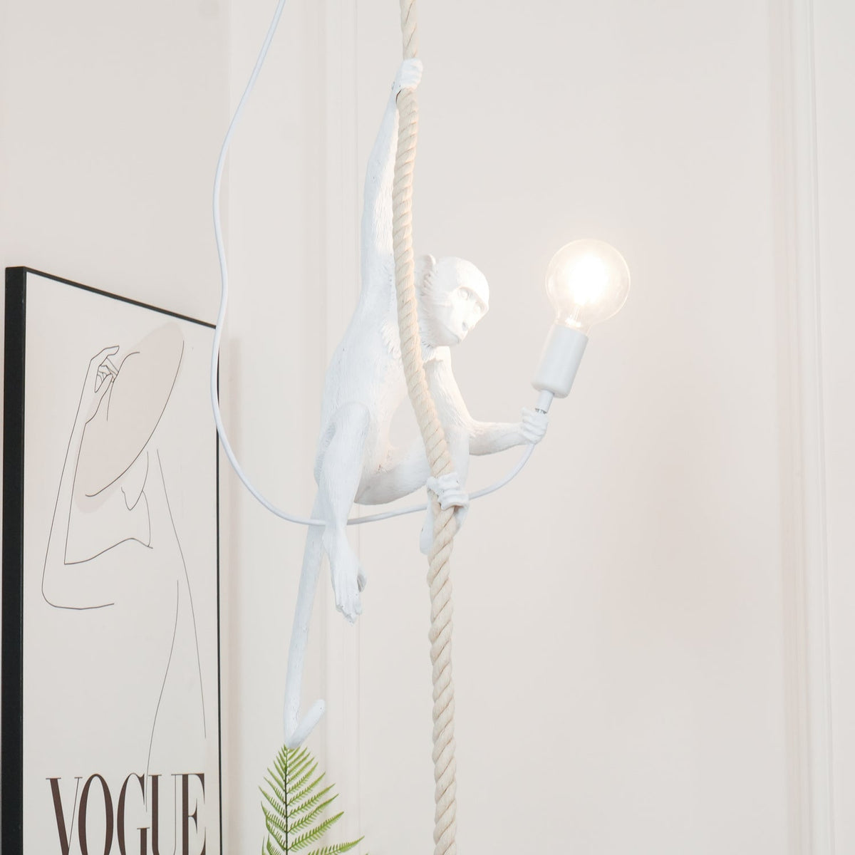 Long-armed white monkey pendant gripping its rope as the round bulb glows, with a framed line drawing partly in view behind it; by Clérachic.
