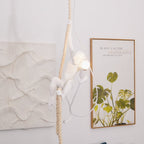 This Clérachic pendant seen from the side, the white monkey clinging to its rope and holding a lit bulb beside a leafy framed print.