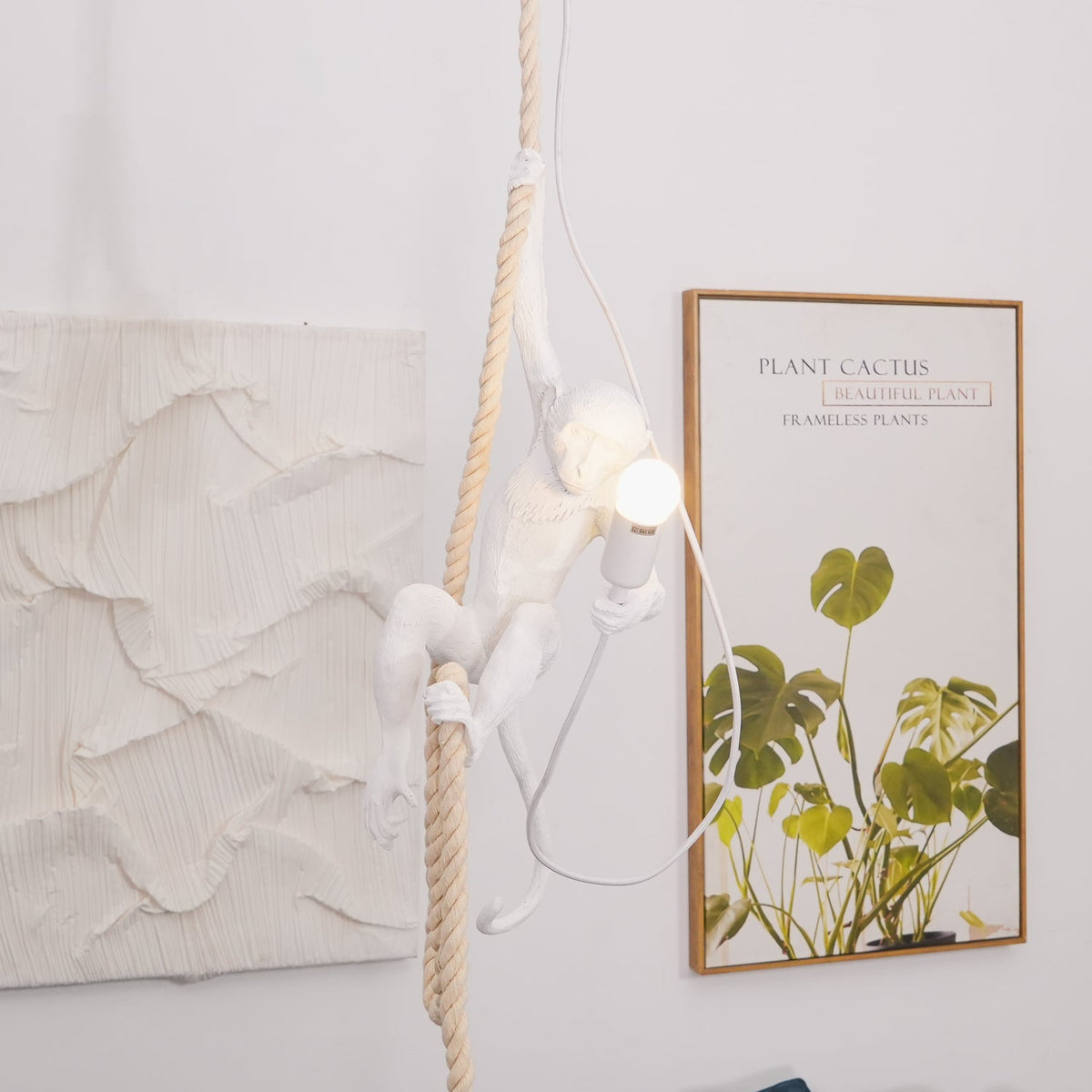 This Clérachic pendant seen from the side, the white monkey clinging to its rope and holding a lit bulb beside a leafy framed print.