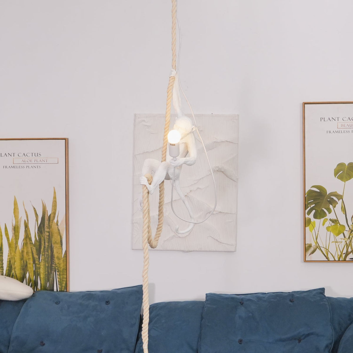 Wide view of a white monkey pendant lit above a blue velvet sofa, with textured plaster art and botanical prints on the pale wall; Clérachic.
