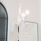 Close three-quarter view of the white monkey on a thick twisted rope, one hand on the bulb holder, beside an arched window; from Clérachic.