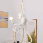 Resin monkey-figure pendant hanging with its long tail curled, one arm stretched toward a bulb, with a sunset painting and a leafy print behind; a Clérachic design.