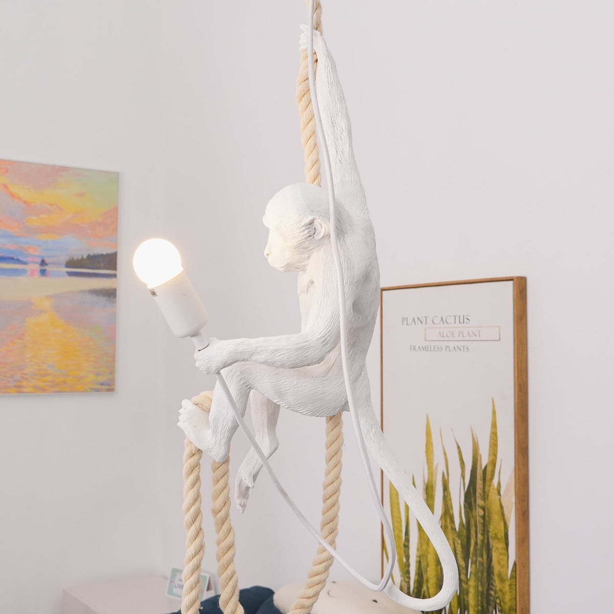 Resin monkey-figure pendant hanging with its long tail curled, one arm stretched toward a bulb, with a sunset painting and a leafy print behind; a Clérachic design.