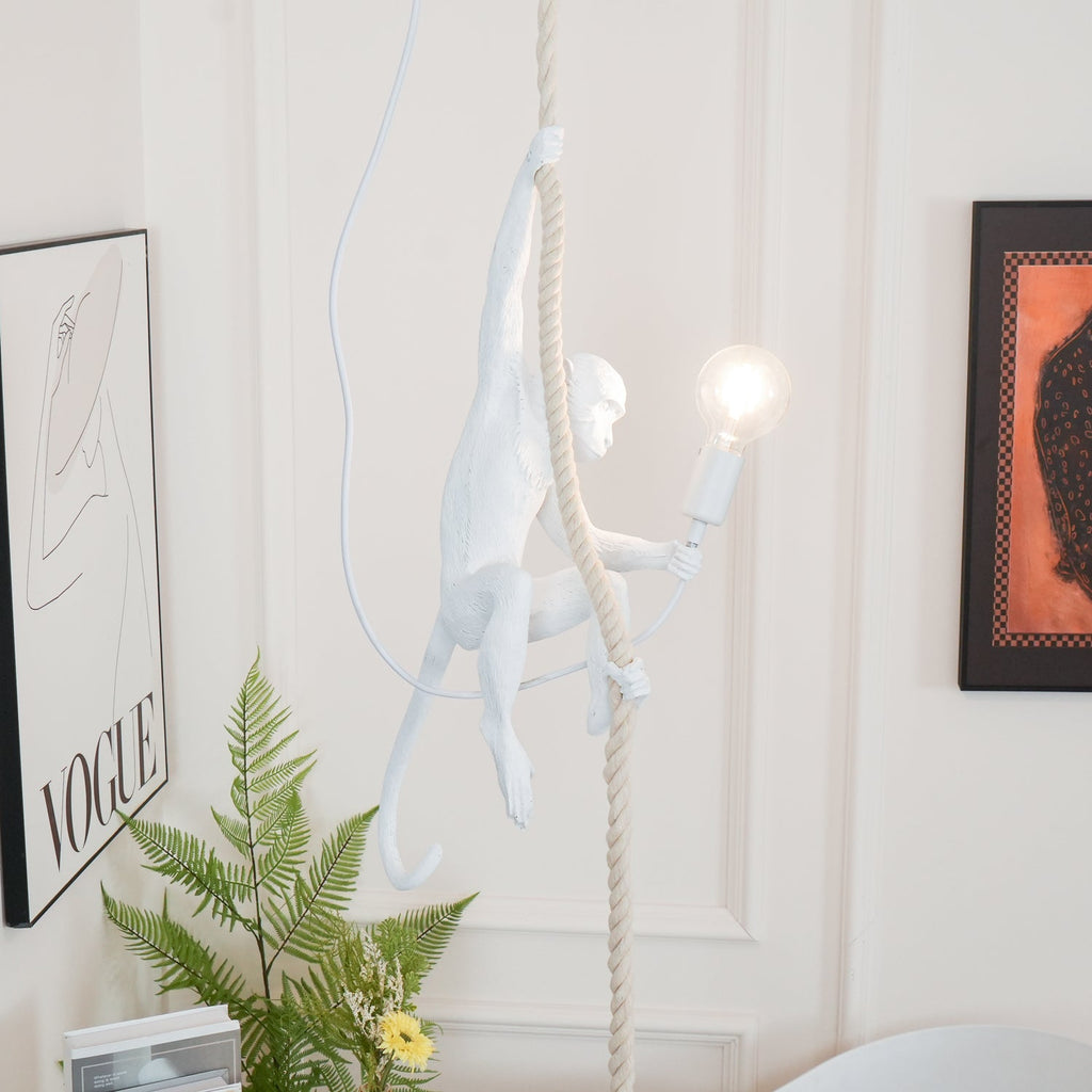 White monkey pendant with its tail curled behind, holding a lit bulb in front of a panelled white wall, a fern and a framed print; from Clérachic.