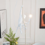 White monkey pendant with its tail curled behind, holding a lit bulb in front of a panelled white wall, a fern and a framed print; from Clérachic.