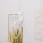 Seated white monkey on a doubled rope, lit bulb beside it, in front of a leafy framed print and textured plaster panel; Clérachic pendant.