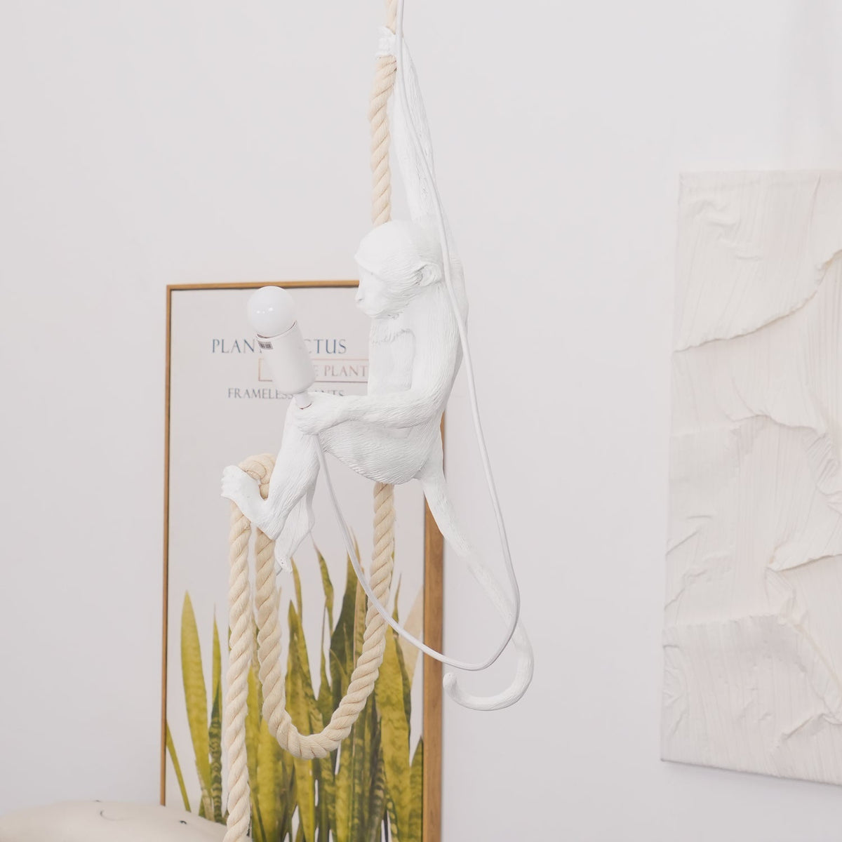 Seated white monkey on a doubled rope, lit bulb beside it, in front of a leafy framed print and textured plaster panel; Clérachic pendant.