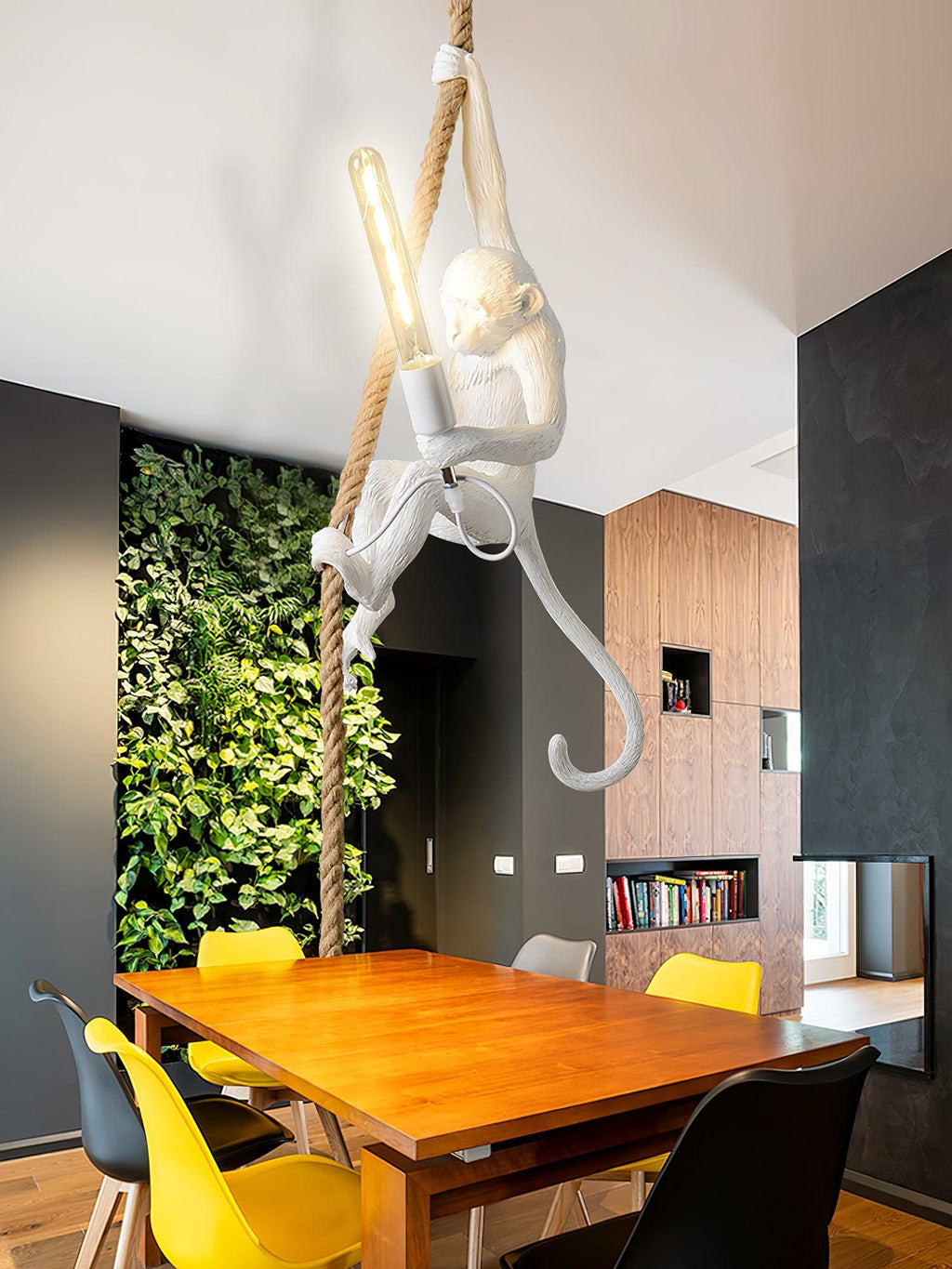 White Clérachic monkey pendant with a long filament bulb, hanging over a wooden dining table with yellow and black chairs and a plant wall.
