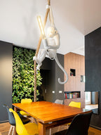 White Clérachic monkey pendant with a long filament bulb, hanging over a wooden dining table with yellow and black chairs and a plant wall.