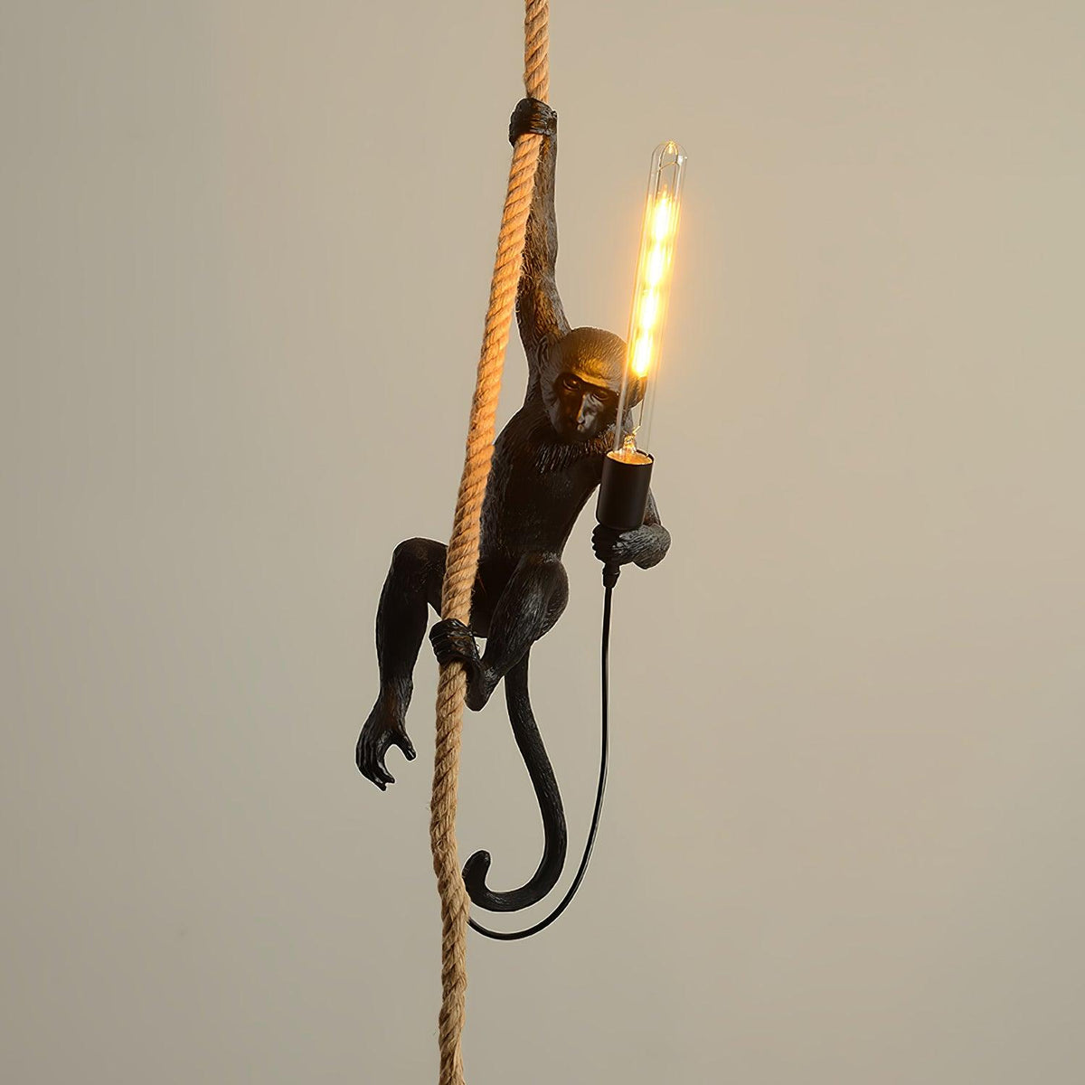 Black monkey on a natural rope holding a long filament bulb that glows amber, against a plain warm beige wall; this Clérachic pendant.
