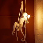 Warm orange light fills a rough brick corner as the white monkey climbs its rope with a glowing bulb in hand; Clérachic pendant.
