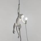 White monkey hanging from a dark cord against a pale grey wall, one hand holding a small bulb, with a dark cable below; a Clérachic pendant.