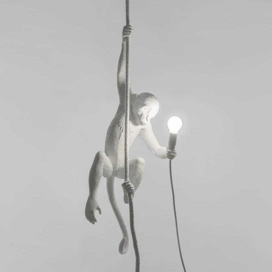White monkey hanging from a dark cord against a pale grey wall, one hand holding a small bulb, with a dark cable below; a Clérachic pendant.