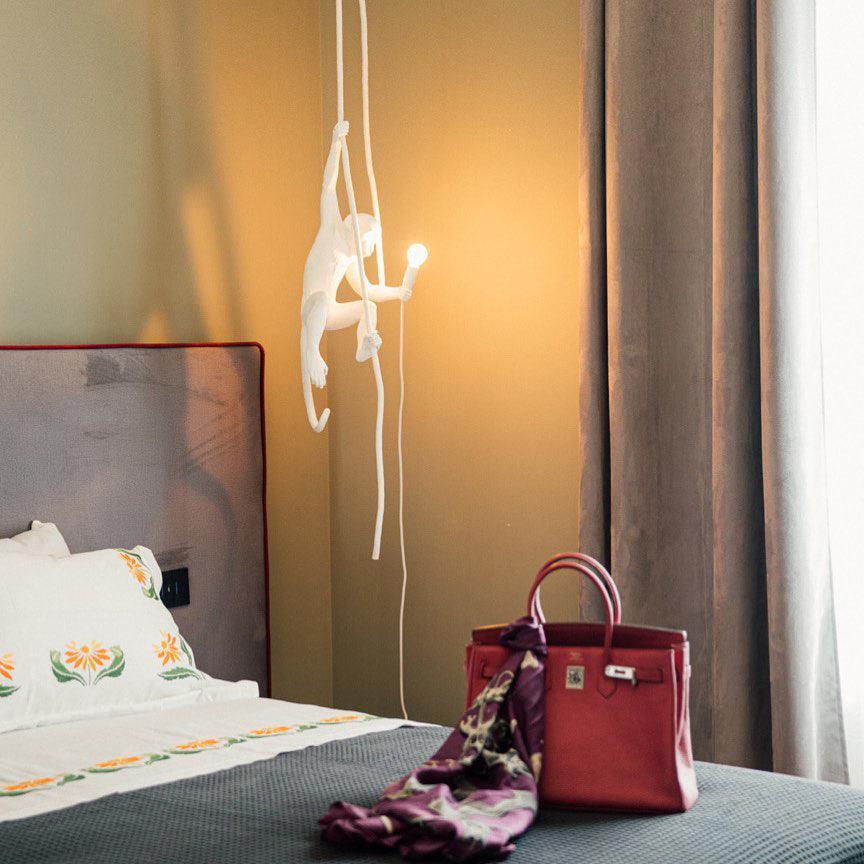 Small white monkey pendant glowing beside a bed with a grey cover and warm-toned wall, a red handbag and scarf at the bottom; from Clérachic.