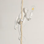 A white Clérachic monkey climbs a rope holding a clear filament bulb in a socket, shown against a pale beige wall with its cord looped below.