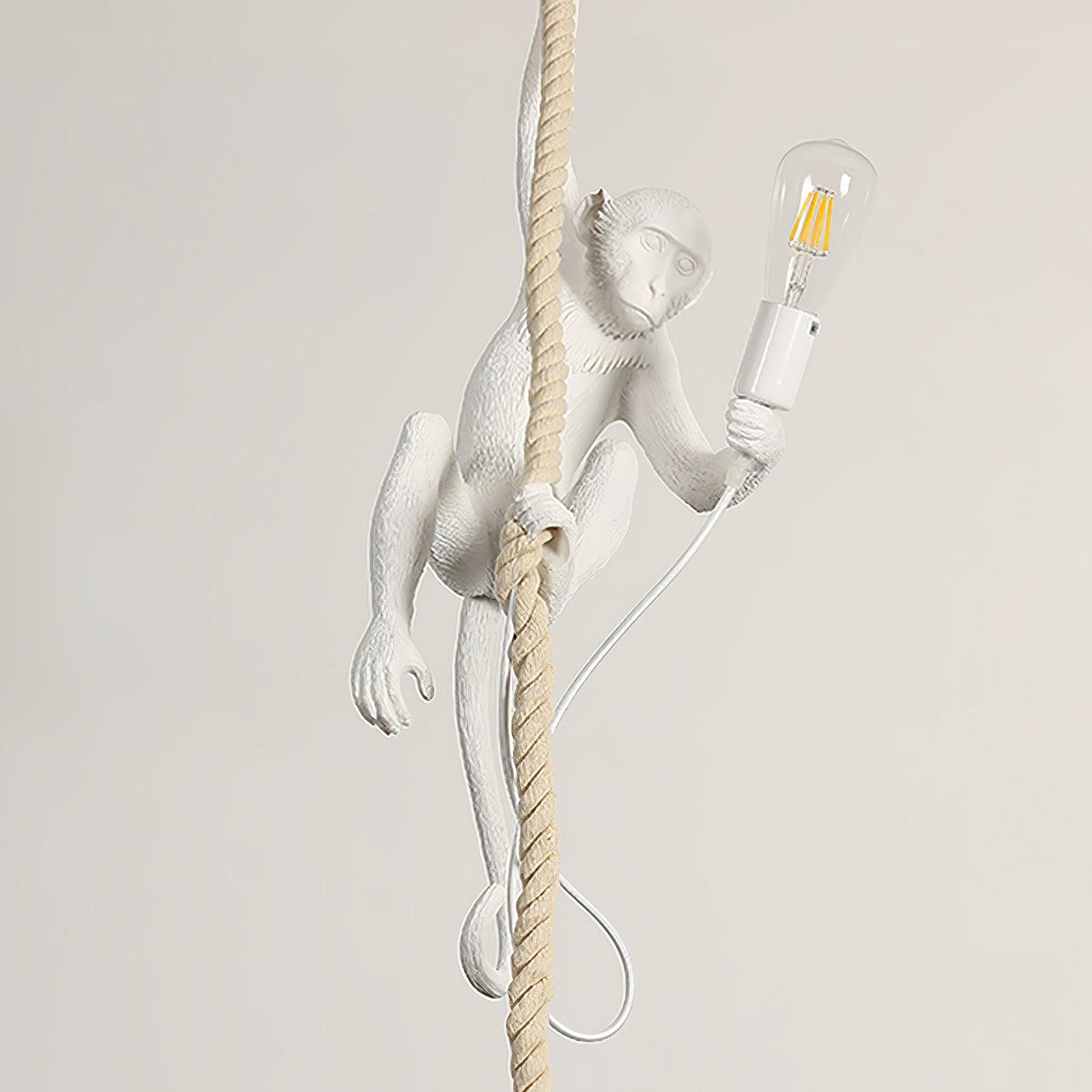 A white Clérachic monkey climbs a rope holding a clear filament bulb in a socket, shown against a pale beige wall with its cord looped below.