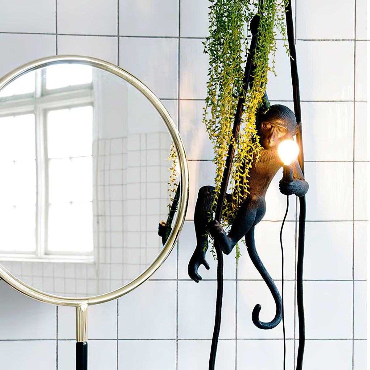 Black monkey pendant with a glowing bulb, trailing greenery wound around its rope, beside a round brass mirror and a white-tiled wall; Clérachic.