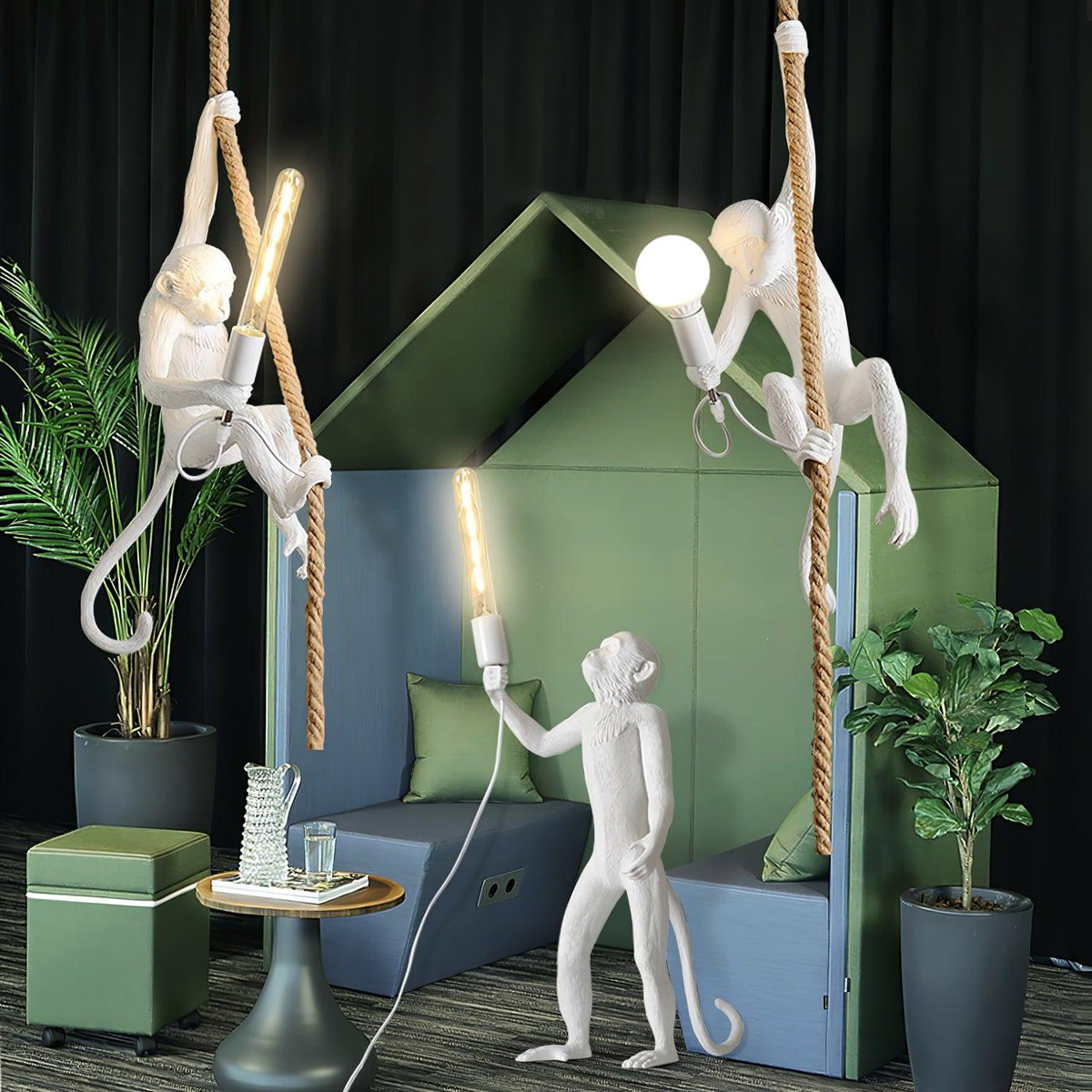 Three white monkey figures in a green and blue booth, two hanging from ropes with lit bulbs and one standing, in a dark room; from Clérachic.