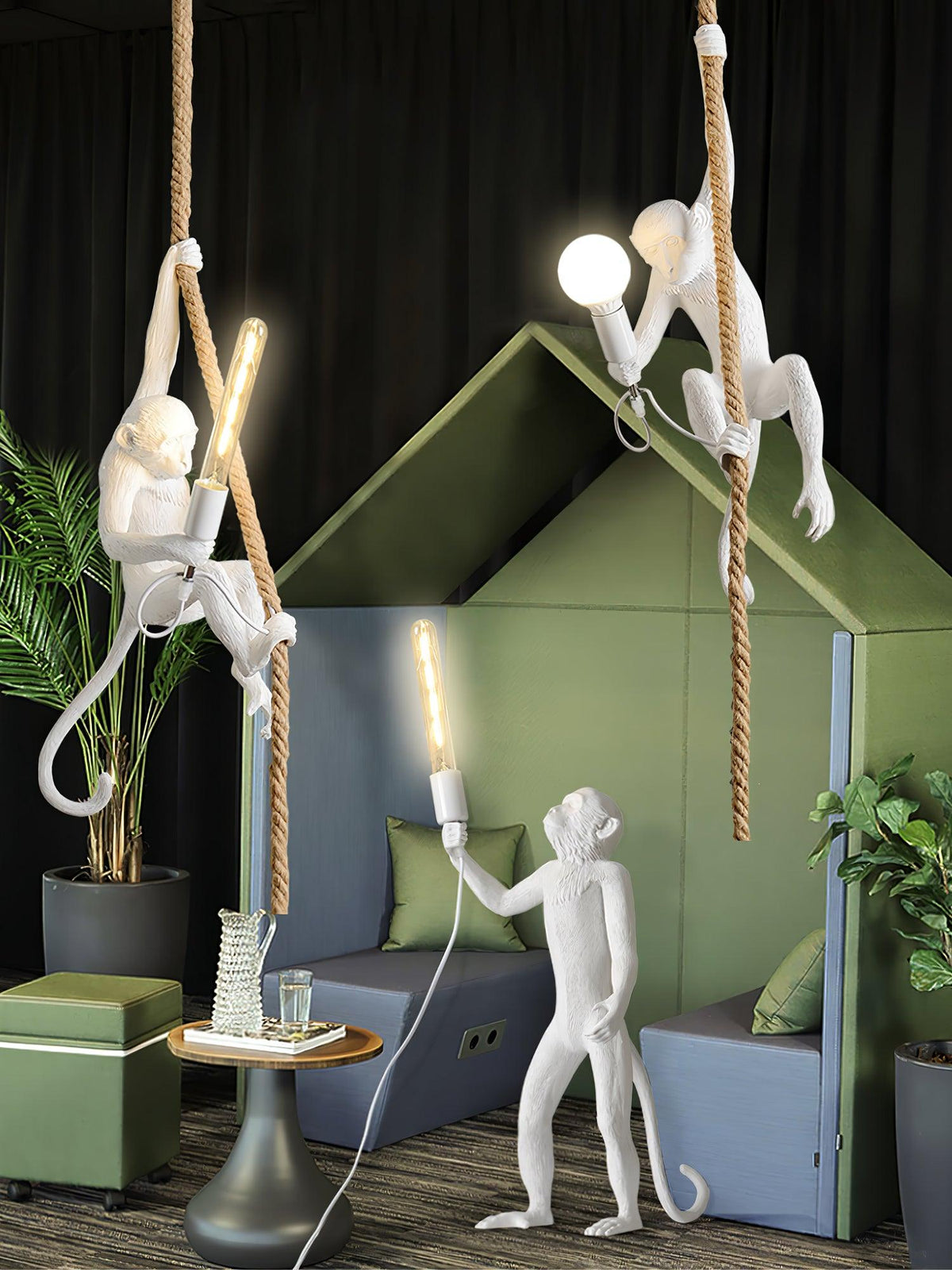 White monkey pendants with lit bulbs beside a standing monkey light, set against a green house-shaped screen and dark backdrop; from Clérachic.