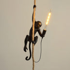 Black monkey on a thick rope holding a long glowing filament bulb, against a plain beige wall, as a Clérachic pendant.