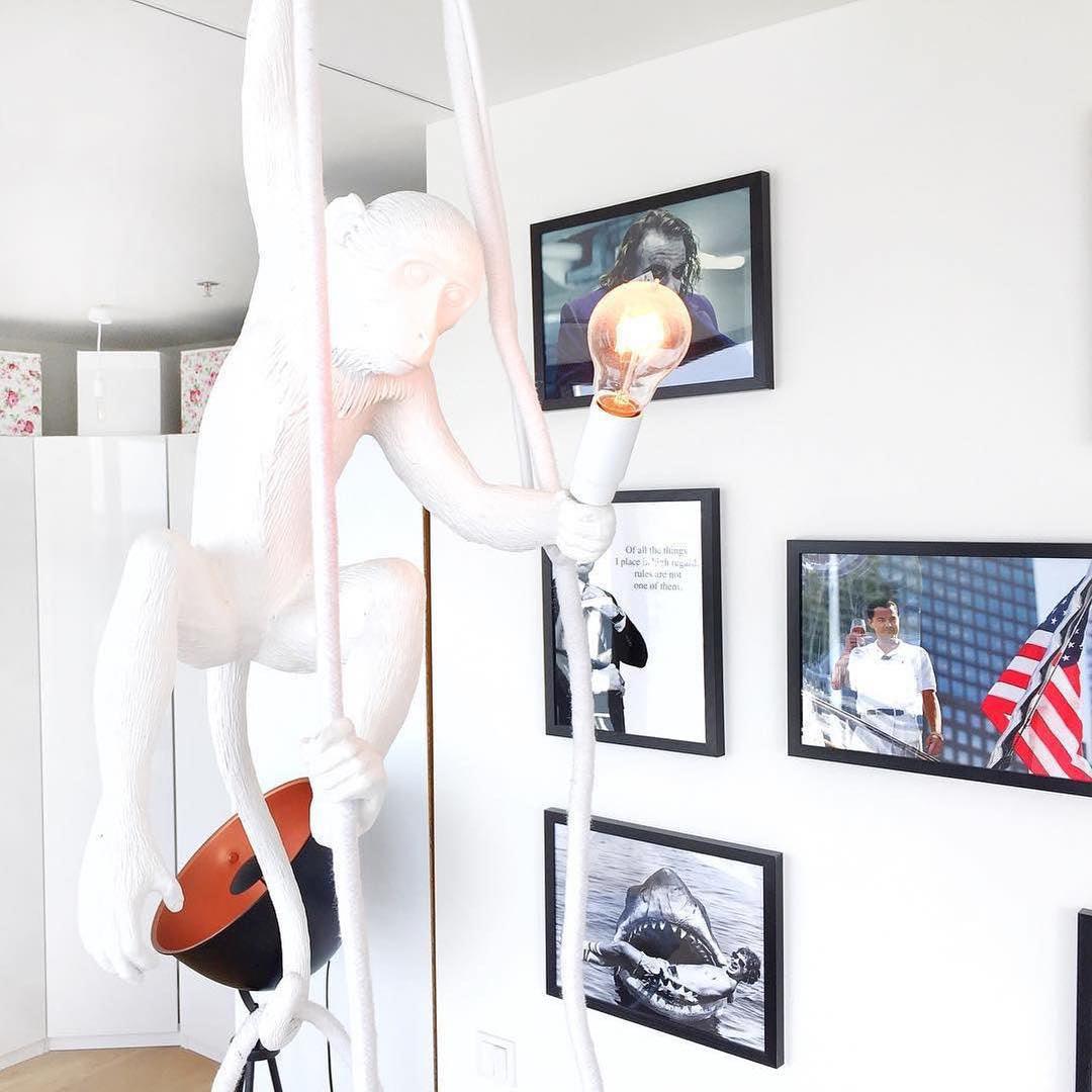 Close view of the white monkey's arm and lit bulb with framed black-and-white photographs and a dark copper-lined lamp behind; Clérachic.