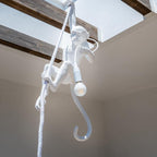 White monkey pendant hanging from a skylight in a beamed ceiling, its rope wrapped by one arm and a round bulb glowing; from Clérachic.