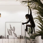 Black monkey pendant in silhouette beside a large palm leaf, holding a small round bulb on a dark cord, in a bright room; this Clérachic design.