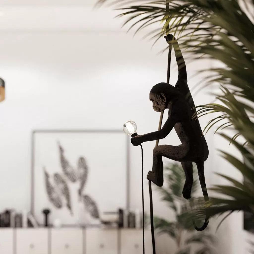 Black monkey pendant in silhouette beside a large palm leaf, holding a small round bulb on a dark cord, in a bright room; this Clérachic design.
