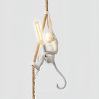 White monkey with one arm stretched up the rope and a long filament bulb lit in its hand, on a pale background; this Clérachic pendant.