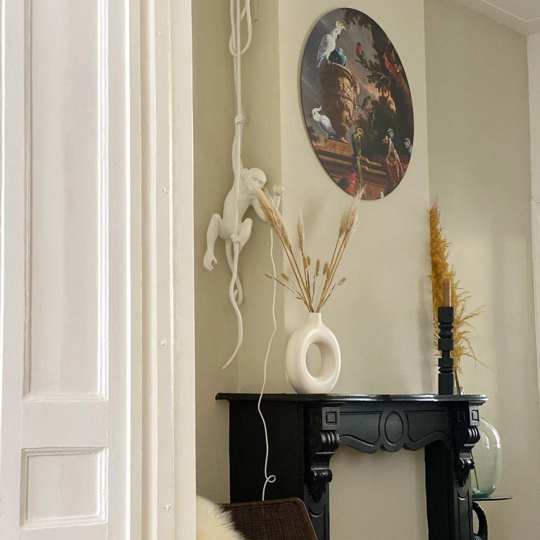 White monkey pendant hung in a hallway beside a black fireplace with a round painting above, dried grasses in a vase below; from Clérachic.