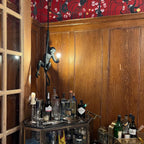 Black monkey pendant with a glowing bulb above a brass drinks trolley, against wood panelling and red patterned wallpaper; a Clérachic piece.