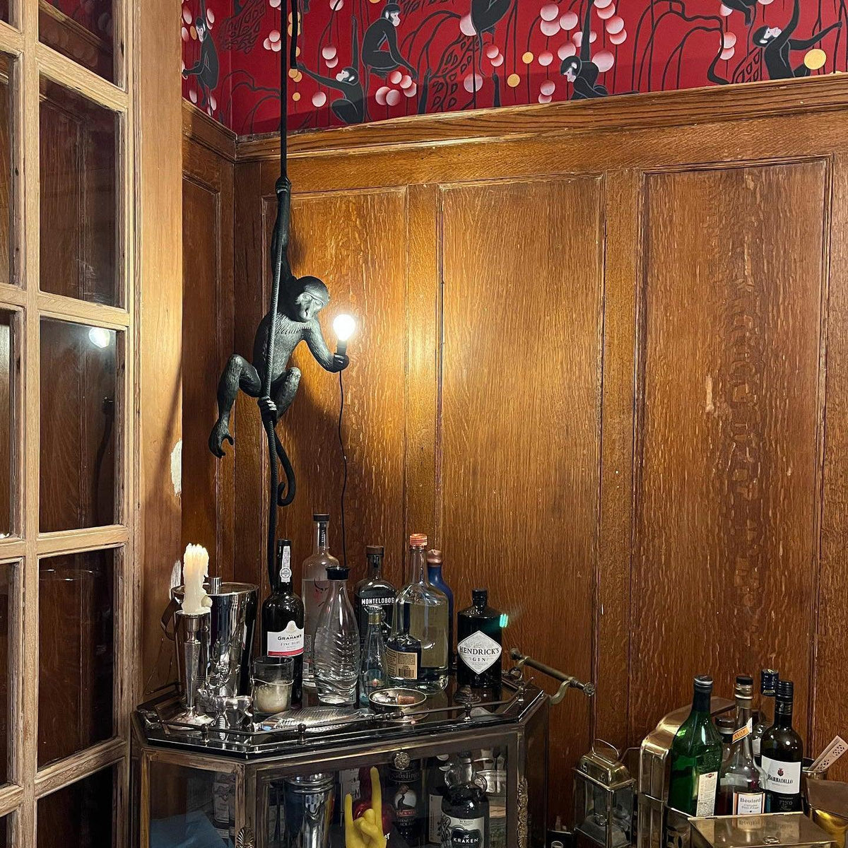 Black monkey pendant with a glowing bulb above a brass drinks trolley, against wood panelling and red patterned wallpaper; a Clérachic piece.