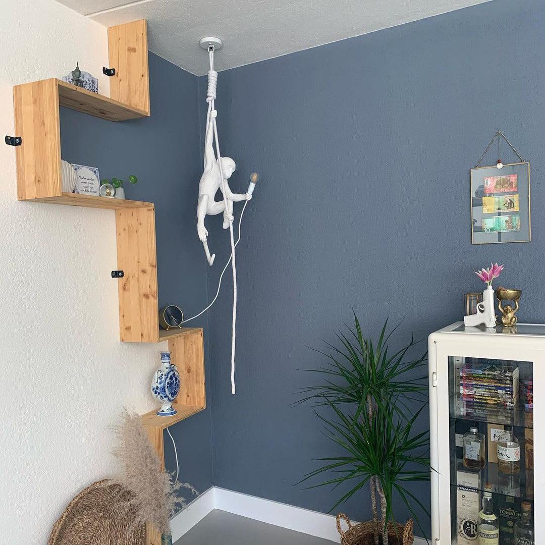 This Clérachic pendant hangs from a white ceiling canopy in a blue room with pale wood shelves, the white monkey holding its lit bulb.