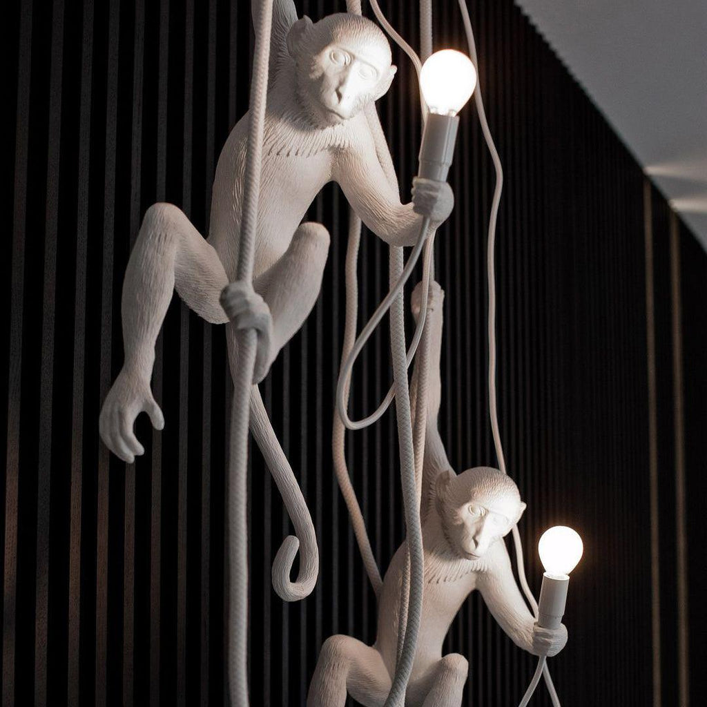 Two white monkey pendants on ropes, both holding lit bulbs, in front of a black slatted wall; a close view of the Clérachic design.