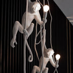 Two white monkey pendants on ropes, both holding lit bulbs, in front of a black slatted wall; a close view of the Clérachic design.