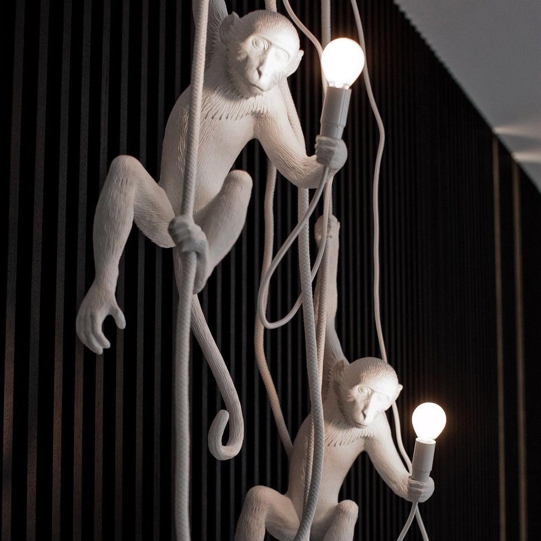 Two white monkey pendants on ropes, both holding lit bulbs, in front of a black slatted wall; a close view of the Clérachic design.