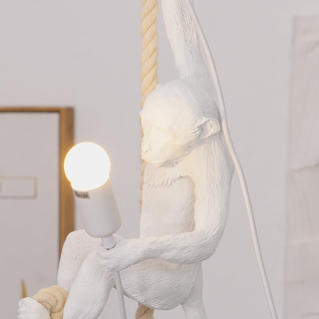 Cropped view of the white monkey's body and a lit round bulb on this Clérachic pendant, with thick rope at the top and a pale wall behind.