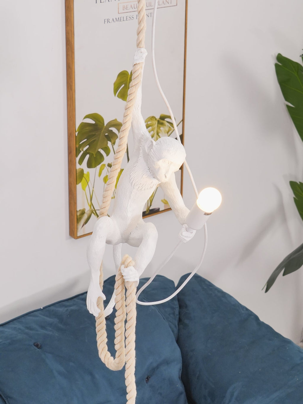 White monkey pendant swinging low with a doubled rope above a blue velvet cushion, a leafy print behind it; from Clérachic.