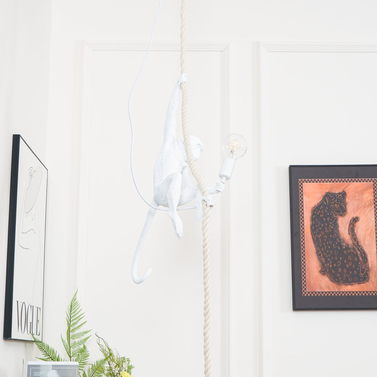 Distant view of the white monkey pendant on a slim rope against a panelled wall, with a leopard print and a fern; this Clérachic design.