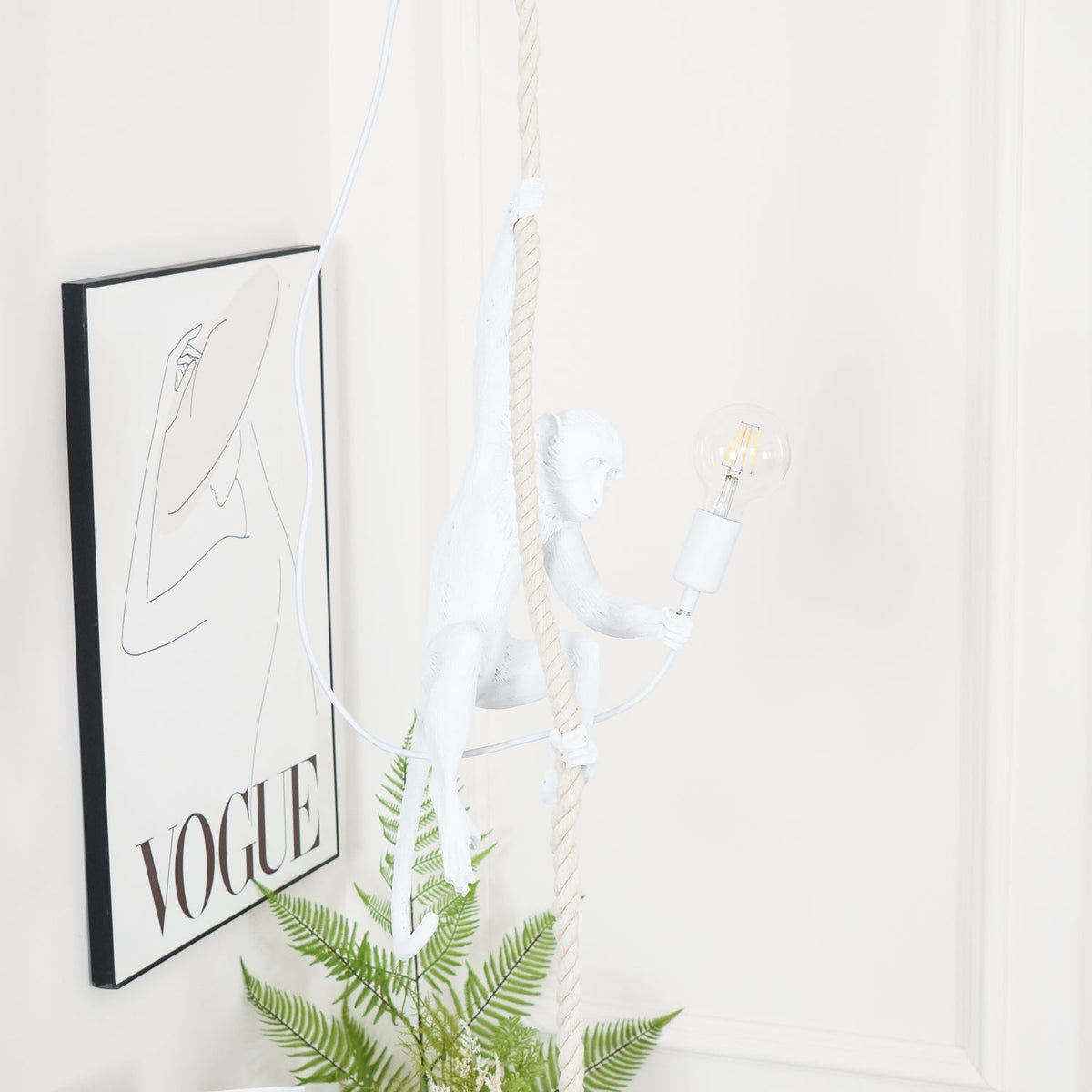 This Clérachic pendant frame shows the white monkey seated low on its rope, a filament bulb lit, beside a black-framed print and a fern.