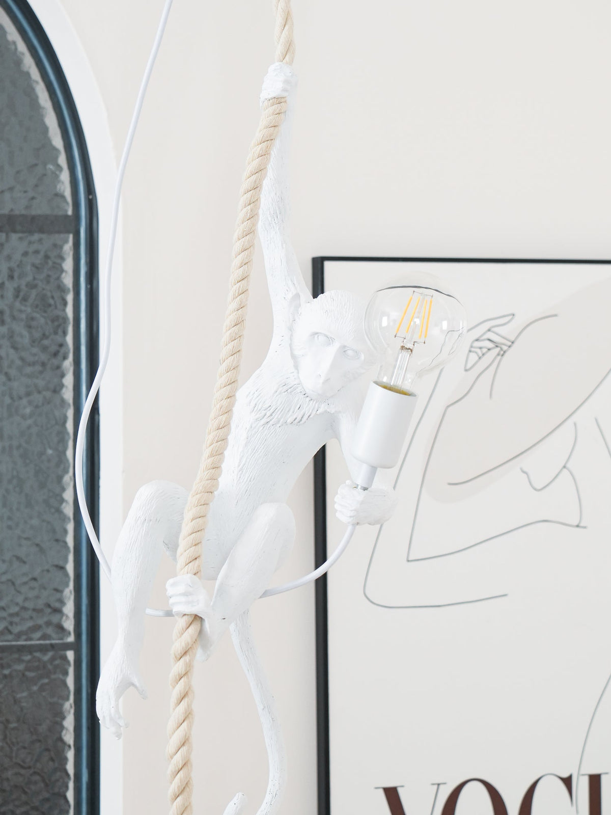 Tall view of the white monkey on a thick rope with its arm up and a bulb glowing, beside an arched window and a line-drawing print; Clérachic.