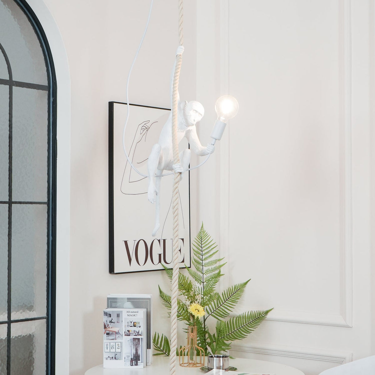 White monkey with a glowing bulb hanging above a table with a fern and magazines, in front of an arched window; from Clérachic.