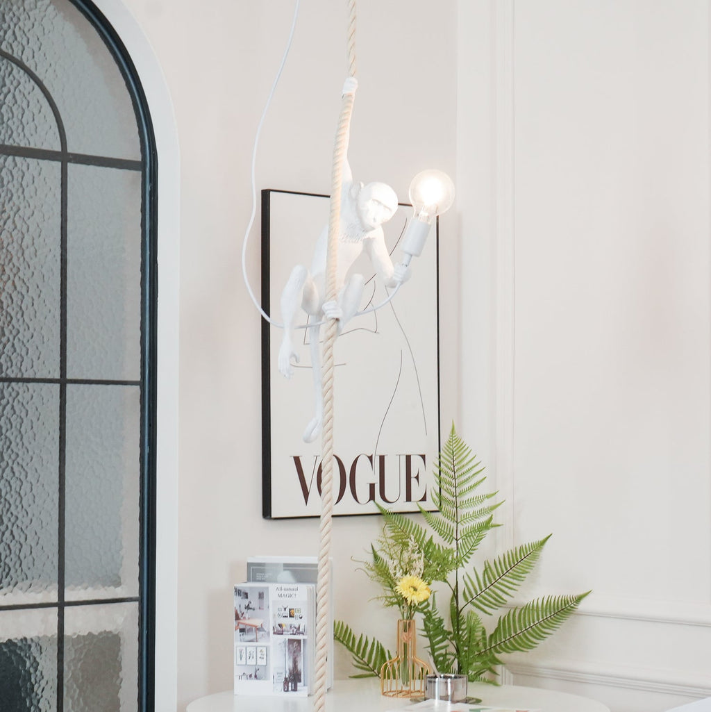 Distant view of the white monkey pendant lit beside a black-framed print and an arched window, a fern and flowers below; Clérachic.