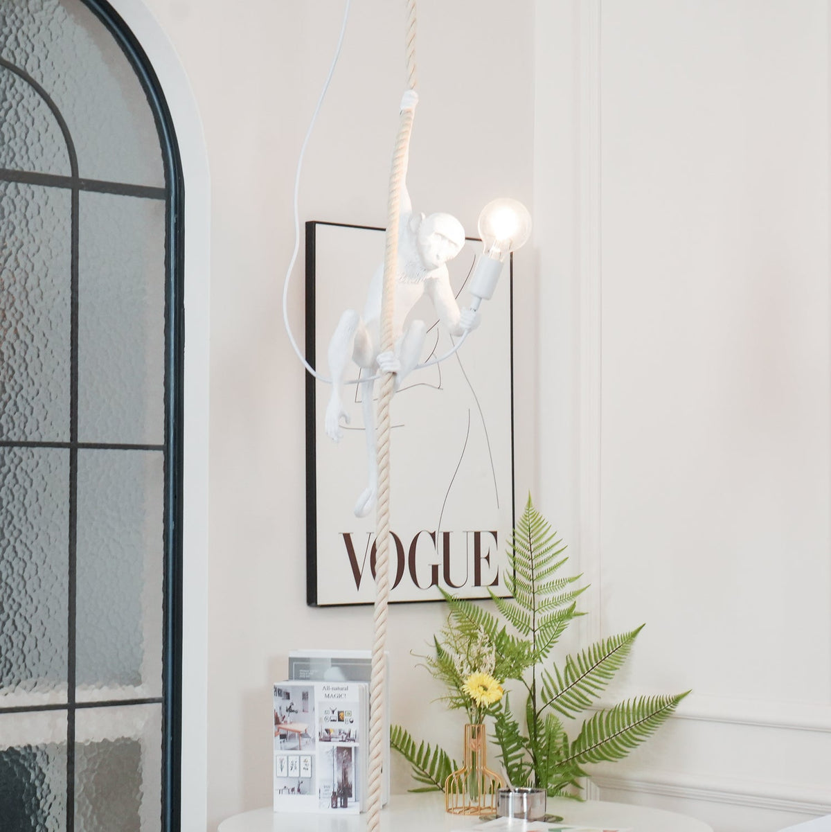 Distant view of the white monkey pendant lit beside a black-framed print and an arched window, a fern and flowers below; Clérachic.