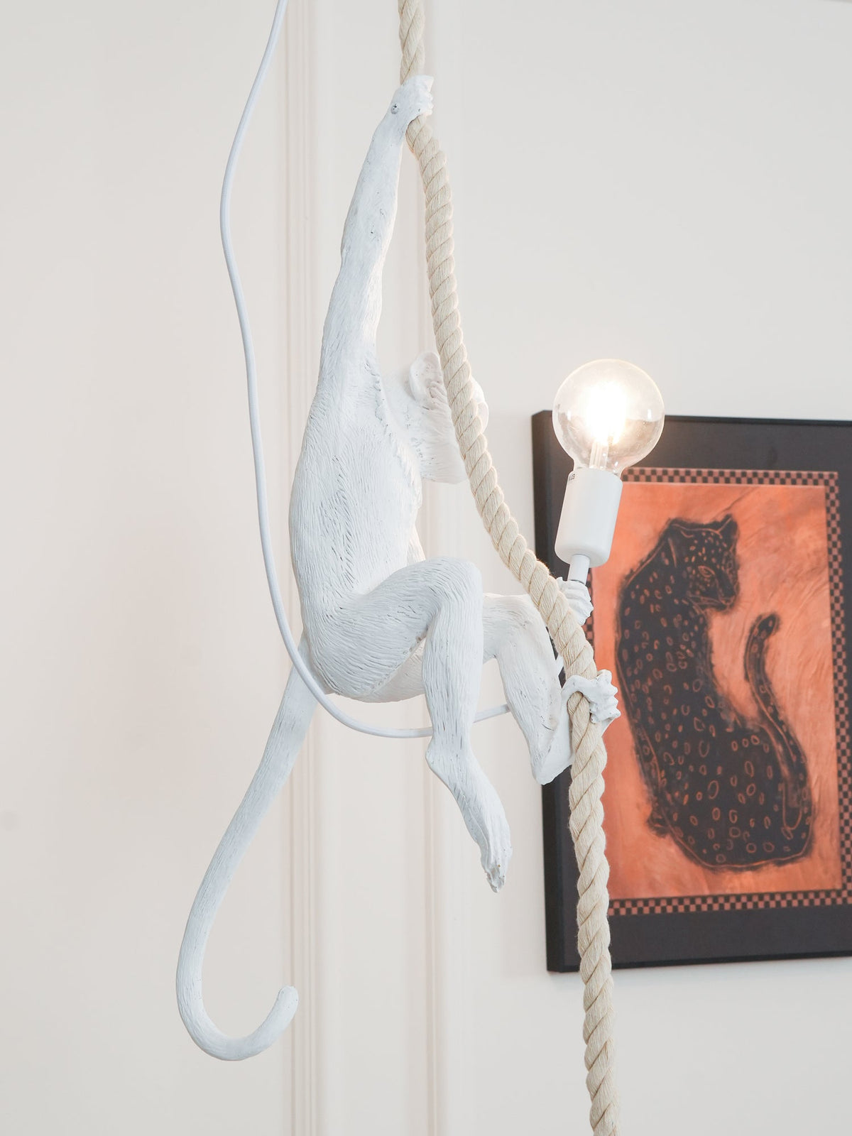 White monkey with its tail curled hanging from a rope, a round bulb lit beside a framed leopard print; this Clérachic pendant.