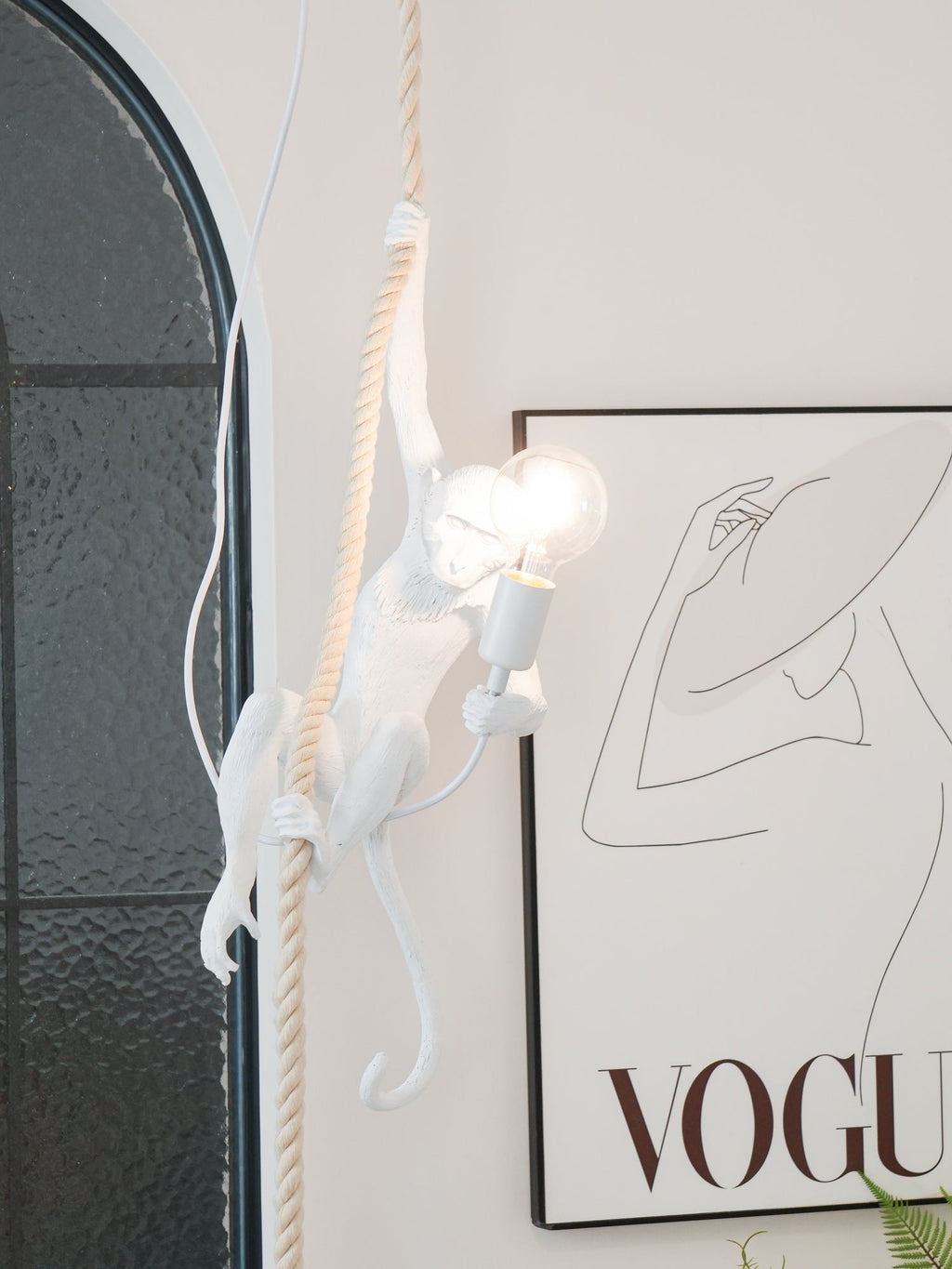 Two bulbs glow beside the white monkey as it climbs its rope, in front of a dark glass window and a large print; this Clérachic pendant.
